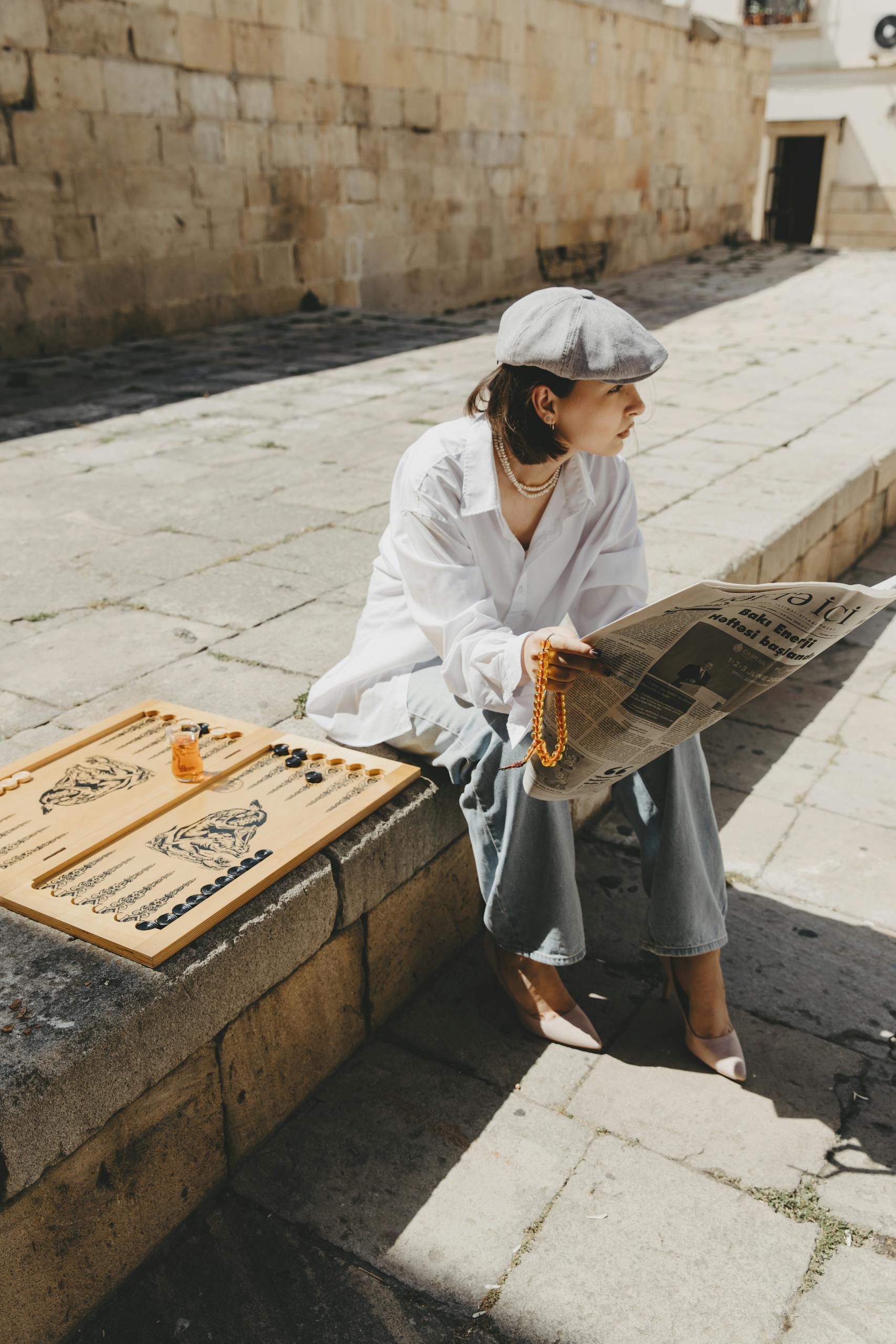 Brunette Woman in Beret and White Shit Sitting on Pavement with Newspaper in Hands