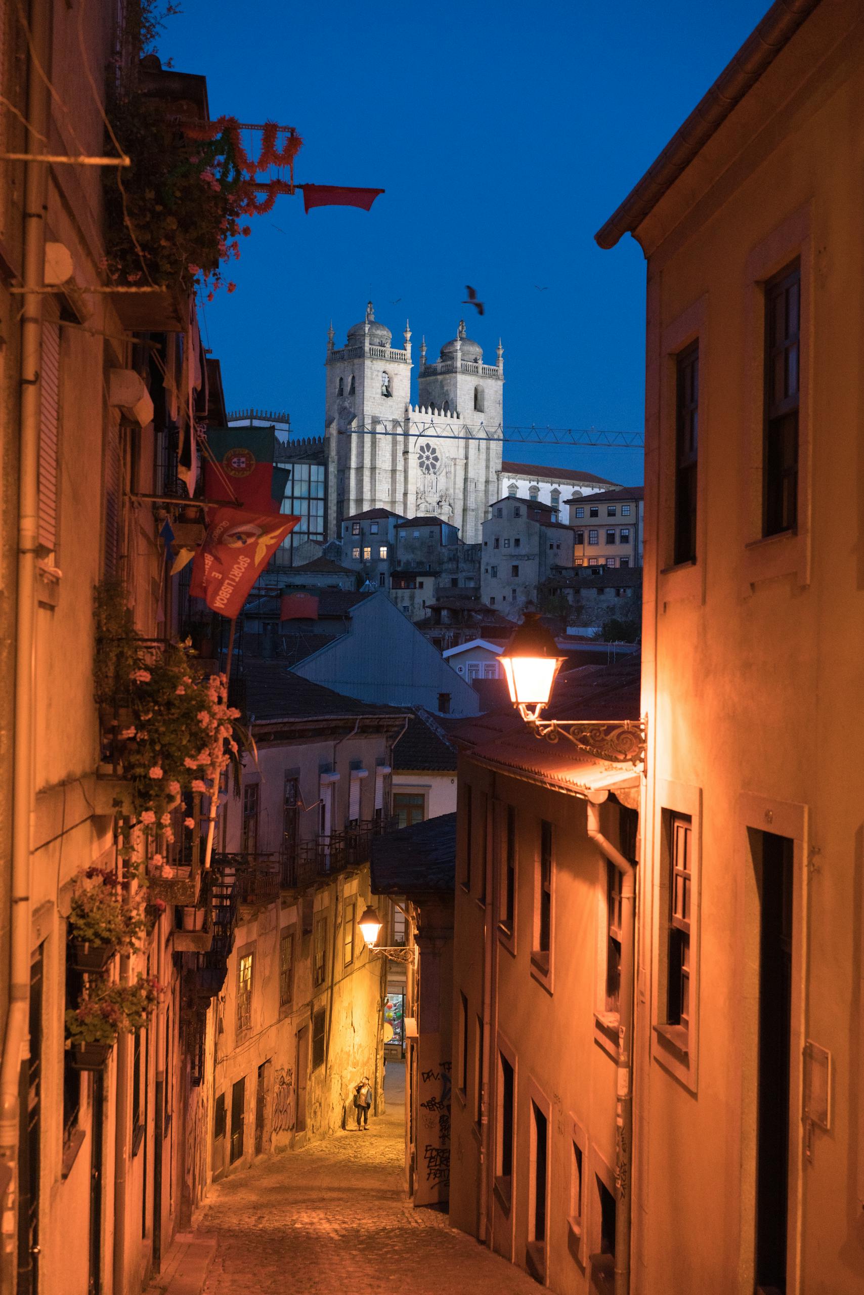 Narrow cobblestone street of Porto City lit with lanterns at dusk with Porto Cathedral on hill in distance
